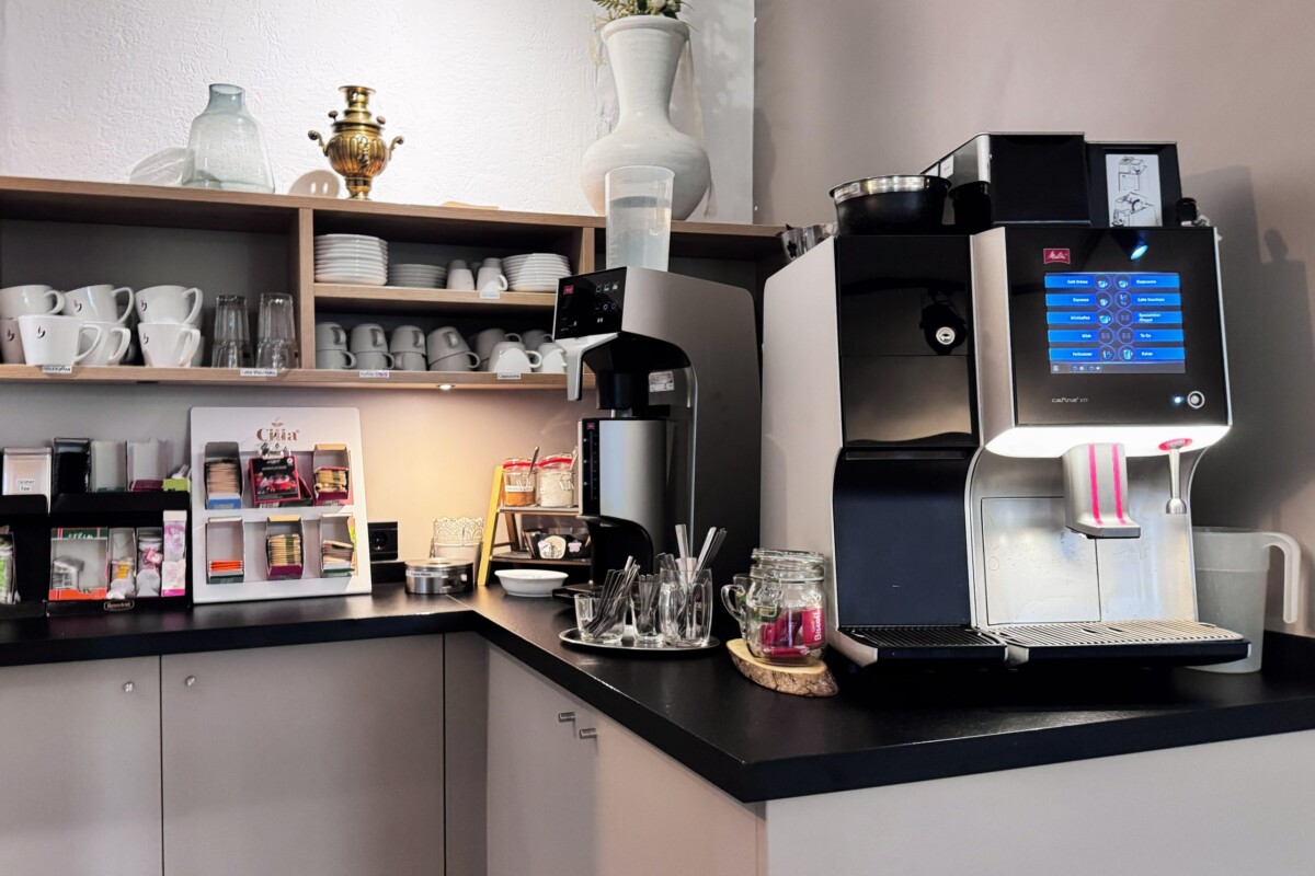 Eine stilvolle Kaffee-Station mit hellbraunem Holzregal, das Weiße Porzellantassen, Gläser und professionelle Barausstattung aufbewahrt. Eine moderne schwarze Espresso-Maschine mit digitalem Touchscreen-Display steht auf der schwarzen Arbeitsplatte. Dekorative Objekte wie eine goldene Vintage-Urne, eine weiße keramische Vase und grüne Topfpflanzen verleihen elegante Akzente. Die moderne, helle Gestaltung kombiniert Funktionalität mit ästhetischem Design und schafft ein einladendes, professionelles Café-Ambiente für Hotelgäste.