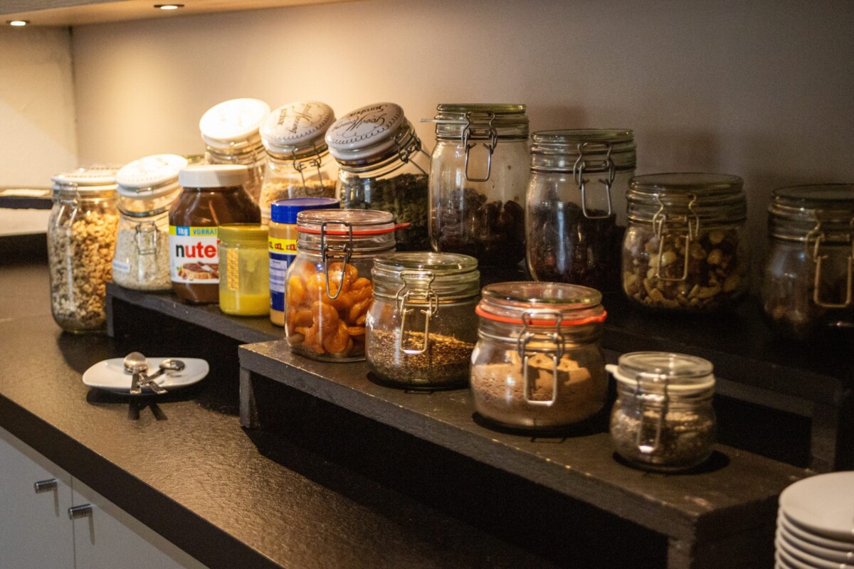 Ein professionell eingerichtetes Café-Buffet-Regal mit schwarzer Arbeitsplatte und warmem Licht. Mehrere transparente Gläser und Behälter mit klappbaren Deckeln präsentieren verschiedene Getreidesorten, Müslis und Cornflakes. Marmeladen, Nutella und andere Fruchtaufstriche stehen in geordneter Anordnung bereit. Weiße Servietten-Halter und Besteck-Organizer vervollständigen das System. Die systematische Anordnung und warme Beleuchtung schaffen ein hygienisches, modernes Frühstücks- und Kaffee-Service-Angebot für gehobene Hotelstandards.