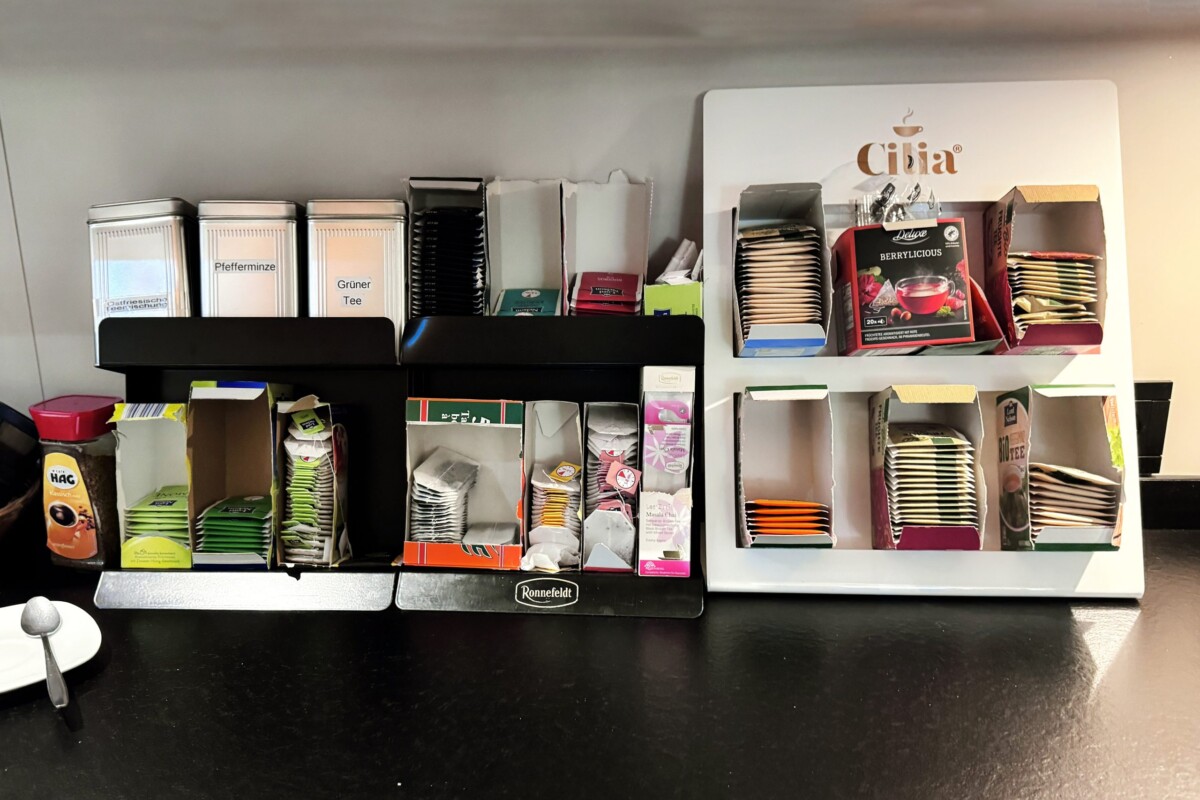 Ein modernes Café-Organisationssystem mit schwarzem Regal präsentiert verschiedene Tee-Sorten, Kaffee-Marken und ein Spendendosen-System. Bunte Boxen in rot, grün und anderen Farben kennzeichnen unterschiedliche Getränke-Kategorien und Zucker-Optionen. Röhrförmige Halter für Zucker-Würfel und Besteck ordnen die Ausstattung übersichtlich. Die professionelle, farblich kodierte Anordnung ermöglicht effizienten Service und Selbstbedienung. Das durchdachte System zeigt gehobene Hotel- und Café-Standards mit praktischer Benutzerfreundlichkeit.