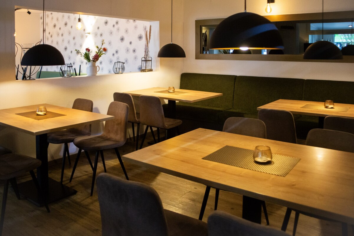 Ein stilvolles Café-Restaurant mit modernem, durchdachtem Design. Mehrere Holztische mit braunen Stühlen bilden Essgruppen. Ein großes samtiges grünes Sofa rahmt die Fensterfront und schafft einen entspannten Loungebereich. Schwarze Pendelleuchten und indirekte warme Beleuchtung schaffen intimates Ambiente. Eine weiße Wand mit graphischem Muster und großem Spiegel vergrößert optisch den Raum. Der Holzfußboden und die durchdachte Möblierung verbinden Komfort mit zeitgenössischem Design und schaffen eine einladende, gehobene Café-Atmosphäre.