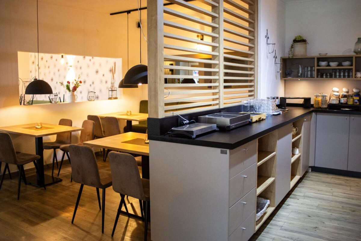 Ein zeitgenössisches Restaurant mit cleverer offener Raumgestaltung: Links befindet sich der Speisebereich mit hellen Holztischen und braunen ergonomischen Stühlen, beleuchtet durch schwarze Pendelleuchten. Eine dekorative weiße Wand mit graphischen Mustern und eine große schwarze Pendelleuchte strukturieren den Raum. Rechts öffnet sich eine moderne Küche mit grauem Küchenblock, schwarzer Arbeitsplatte und hellem Holzregal-System, das Geschirr und Vorräte übersichtlich zeigt. Horizontale Holzlamellen-Sichtschutz und warme gelbe Beleuchtung schaffen eine gemütliche, moderne Gastronomie-Atmosphäre.